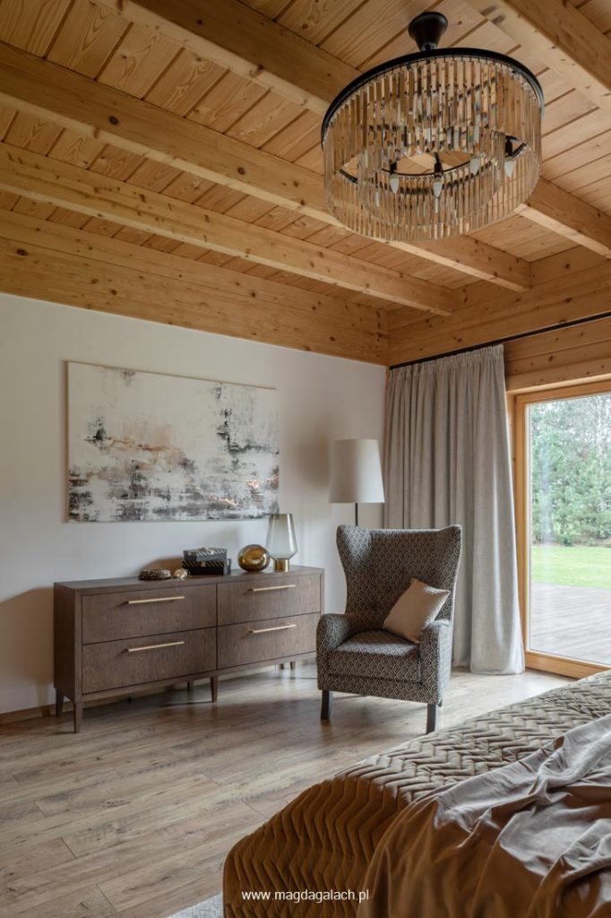 sypialnia w drewnianym domu z drewnianym sufitem, kryształową lampą, drewnianą komodą i fotelem