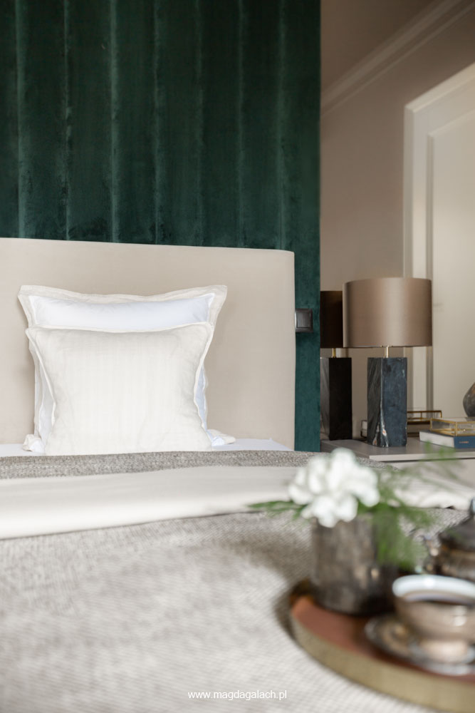 tapicerowane beżowe łóżko z dwoma poduszkami na tle zielonej aksamitnej ściany, obok lampa stołowa