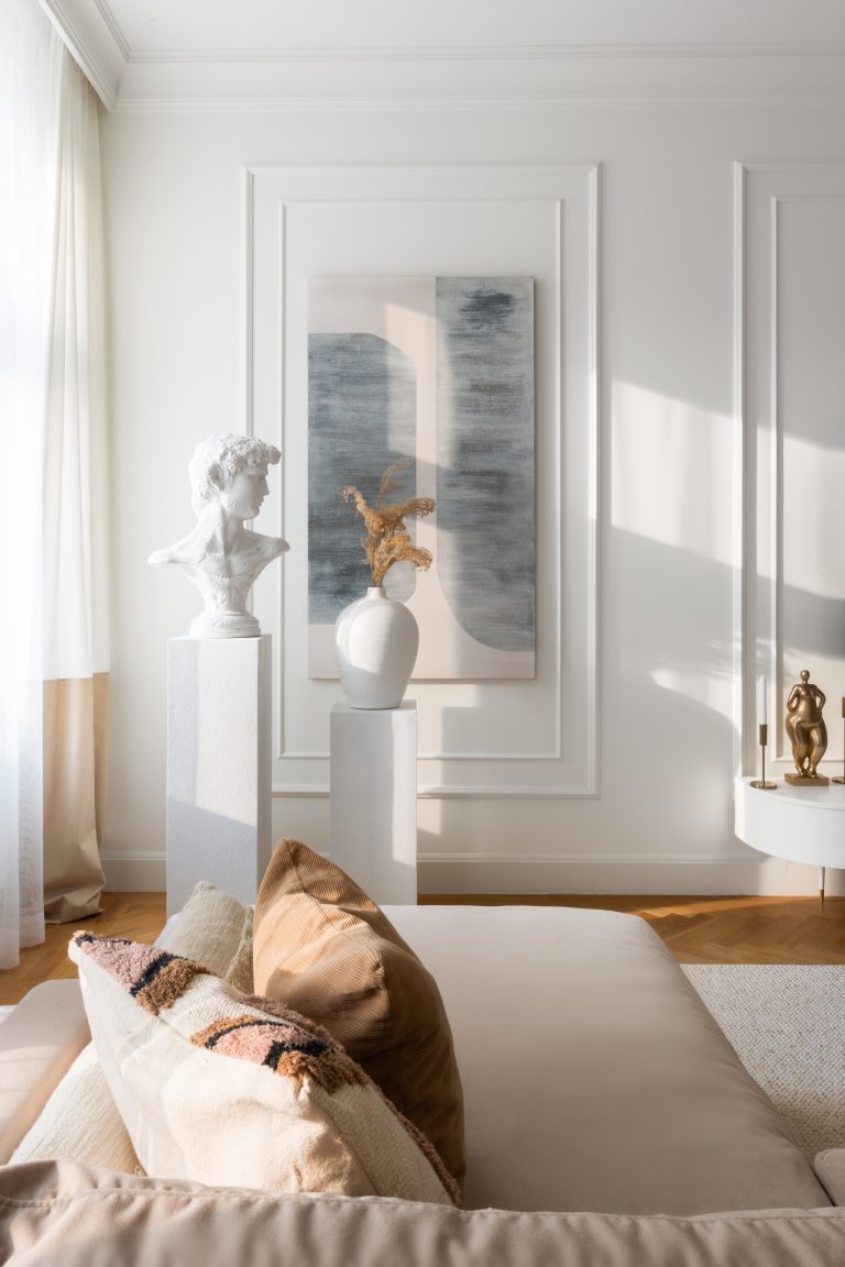 elegancki biały salon ze sztukaterią, rzeźbą i wazonem na postumencie oraz beżową aksamitną kanapa